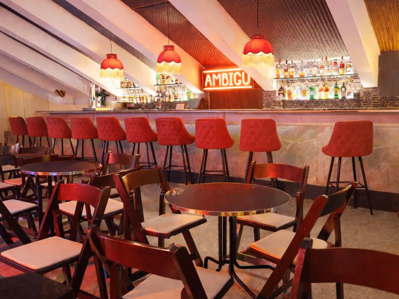Ambigú con barra iluminada en tonos cálidos, taburetes rojos alineados y mesas redondas con sillas en primer plano, ambiente moderno y acogedor.
